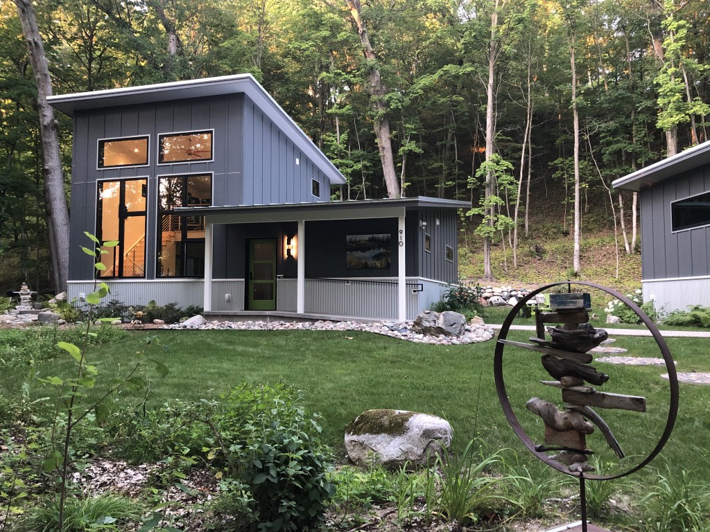 This image shows the Good Hart Artist Residency a modern house in a wooded setting, featuring a clean architectural design with a sloped roof and large windows. The building has a gray-blue exterior with white trim and a green door, adding contrast and character. The landscaping is well-kept, with a neatly mowed lawn, decorative stone paths, and natural-looking gardens. A sculptural art piece made of wood and metal is prominently displayed in the foreground, adding a creative touch to the garden.